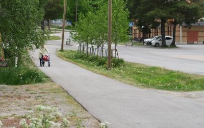 Kuvassa katualuetta Kalannista. Taustalla vanhus rollaattorin kanssa ja autoja parkissa. Pyörätie sekä ajoväylä.